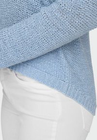 Pull tricoté bleu clair avec motif texturé, porté avec un pantalon blanc ajusté, montrant le côté et le bas du torse de la personne.