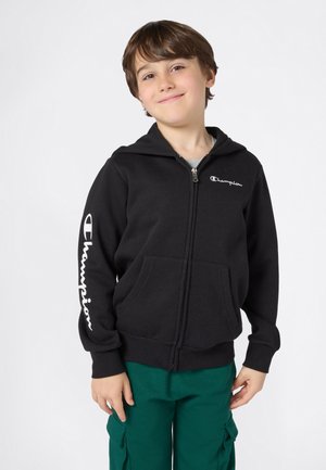 Černá mikina s kapucí na zip vyrobená z bavlny, s přední kapsou; na hrudi a rukávu se nachází bílý logotyp "Champion". Doplněná o zelené kalhoty.