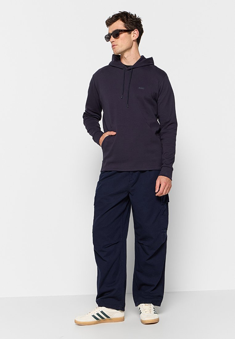 Sweatshirt à capuche bleu marine avec des manches longues et une poche frontale, associé à un pantalon cargo bleu marine ample et des baskets blanches avec des rayures vertes.