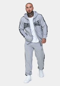 Sivý bavlnený set mikiny a teplákov s predným logom, čiernymi bočnými pruhmi a bielymi teniskami. Model stojí v neutrálnej póze.