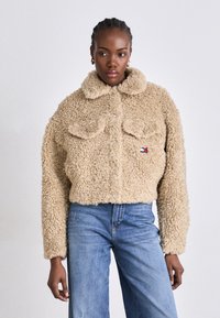 Beige kunstpelsjakke med krage, knappeåpning og to brystlommer. Har et lite logo-patch. Kombinert med lyseblå jeans.