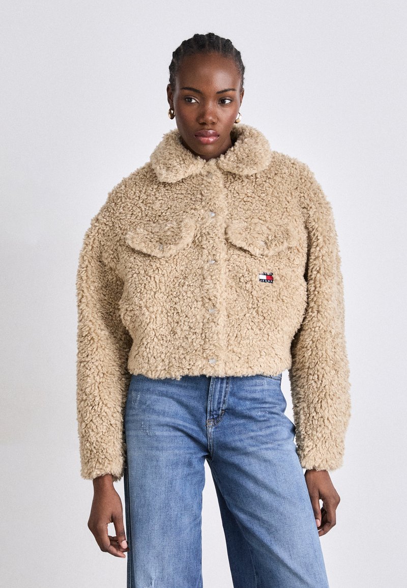 Beige kunstpelsjakke med krage, knappeåpning og to brystlommer. Har et lite logo-patch. Kombinert med lyseblå jeans.