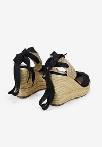 Sandales compensées avec une semelle en jute, des brides en tissu noir et un dessus en beige texturé. Des rubans noirs noués ajoutent une touche de détail aux talons.