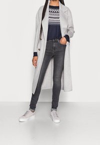 Lungo cappotto grigio sopra un maglione blu a fantasia, abbinato a jeans neri attillati e scarpe da ginnastica platform grigie chiare. Texture liscia, colori neutri.