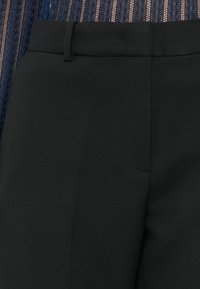 Pantalon noir sur mesure porté avec un haut transparent texturé bleu foncé révélant la taille et les détails des poches avant.