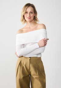 Bijela off-the-shoulder bluzica dugih rukava s drapiranim izrezom, u kombinaciji s visokim bež hlačama s pregibima i prednjim dugmetom.