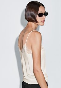 Beige, mouwloos topje met een kantafwerking langs de halslijn. Heeft dunne spaghetti-schouderbandjes en een losse pasvorm, gemaakt van zachte stof. Effen achterkant.