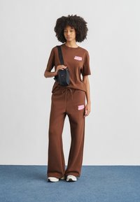 Καφέ βαμβακερό T-shirt με ροζ λογότυπο Nike, σε συνδυασμό με αντίστοιχο φαρδύ παντελόνι και μαύρη τσάντα ώμου. Άσπρα και μαύρα αθλητικά παπούτσια.