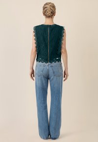 Mujer con el cabello trenzado, vistiendo una blusa sin mangas de encaje verde oscuro con cremallera trasera y jeans de pierna recta azul claro, de pie contra un fondo beige.