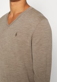 Bežový svetr s výstřihem do V, pleten�á textura, s malým vyšitým logem na levé straně hrudi, úzký střih.