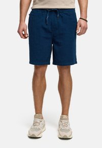Shorts en denim bleu avec une taille élastique et un cordon de serrage. Présentent une texture lisse et des ourlets repliés. Portés avec des baskets de couleur claire.