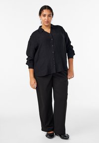 Melnā pogājama krekls ar teksturētu audumu, brīva piegriezuma un garām piedurknēm, kopā ar melnām plaša piegriezuma biksēm un rotātām melnām kurpēm.