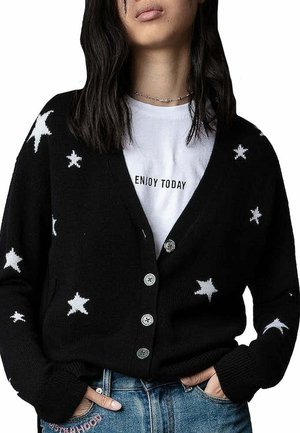 Gilet en tricot noir avec des motifs d'étoiles blanches, décolleté en V et boutons argentés. Porté par-dessus un t-shirt blanc avec du texte imprimé.