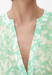 Primo piano del collo e della parte superiore del torace di una persona che indossa una blusa a V di colore verde menta e bianca con un motivo.