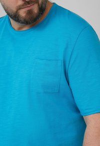 Teal kortemouwen t-shirt van zachte stof, met een borstzak aan de linkerzijde en een ronde halslijn, met een subtiele textuur door het hele shirt.