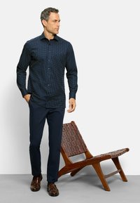 Camicia blu navy con pois bianchi, maniche lunghe, abbinata a pantaloni navy e mocassini marroni, accanto a una sedia in legno intrecciata.