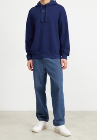 Námorně modrá mikina s kapucí a šňůrkami, doplněná logem, v kombinaci se světle modrými džíny a bílými teniskami. Casual střih a materiály.
