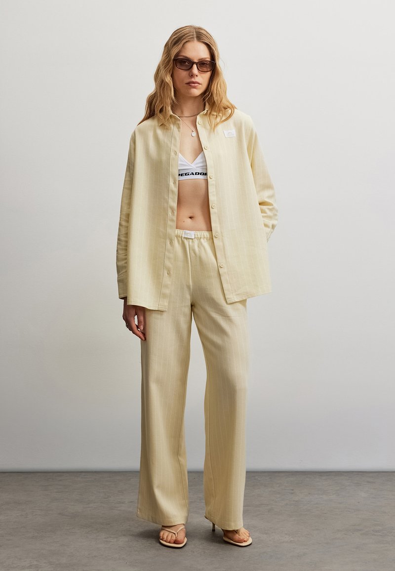Femme aux longs cheveux ondulés portant une chemise beige oversize boutonnée et un pantalon, un soutien-gorge de sport blanc, des lunettes de soleil et des sandales à talons, debout à l'intérieur.
