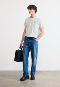 Νεαρός άνδρας που φοράει λευκό T-shirt Calvin Klein, μπλε τζιν, μαύρη ζώνη, μαύρα παπούτσια, κρατώντας μαύρη τσάντα, στέκεται μπροστά σε απλό φόντο.