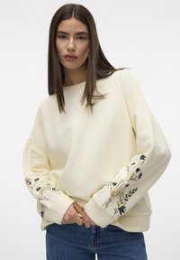 Sudadera crema de manga larga; presenta bordados florales en azul y amarillo en los puños; corte relajado y textura suave.