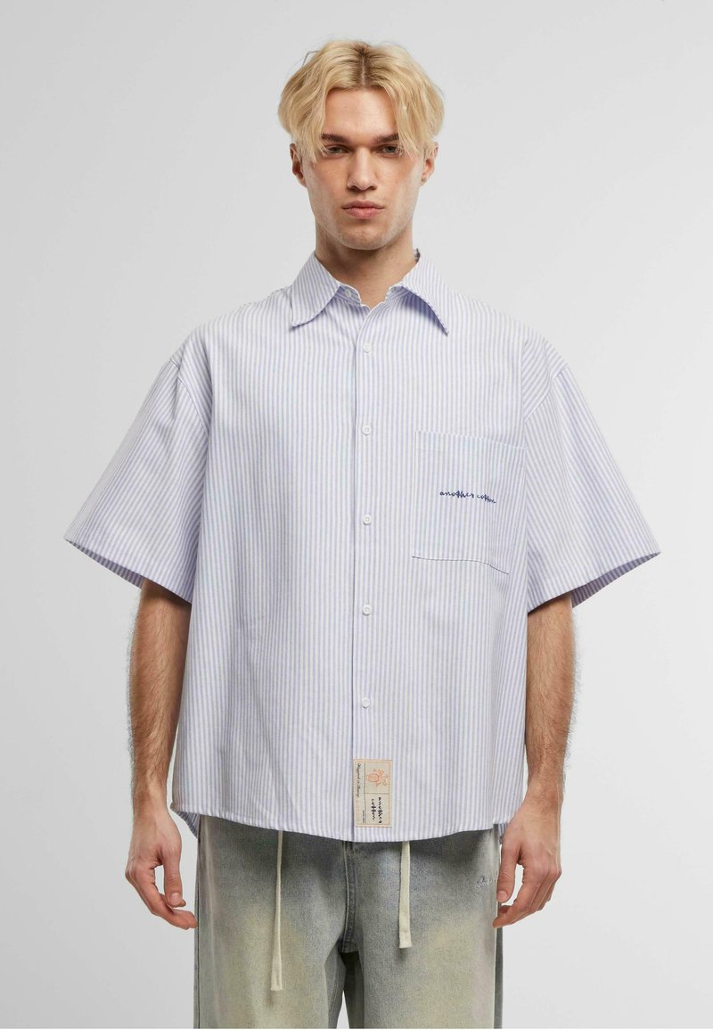 Camicetta a maniche corte a righe in blu chiaro e bianco. Presenta una tasca sul petto con del testo, una vestibilità comoda e un'etichetta tessuta all'orlo.