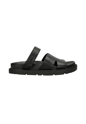 Black leather sandal featuring crossover straps and a flat rubber sole with ridged detailing, offering a modern, minimalist design.