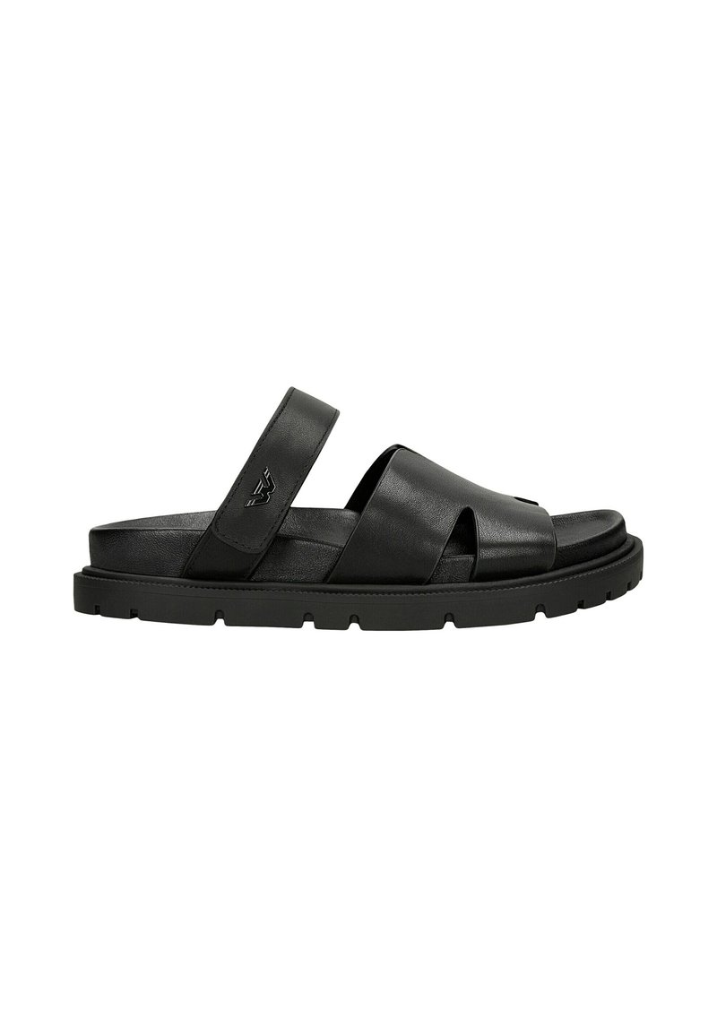 Sandale en cuir noir avec des brides croisées et une semelle en caoutchouc plate avec des détails striés, offrant un design moderne et minimaliste.