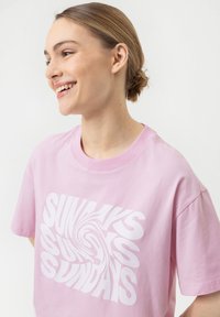 T-shirt en coton rose à manches courtes avec un graphique en texte blanc en tourbillon affichant "DIMANCHES" en évidence sur le devant.