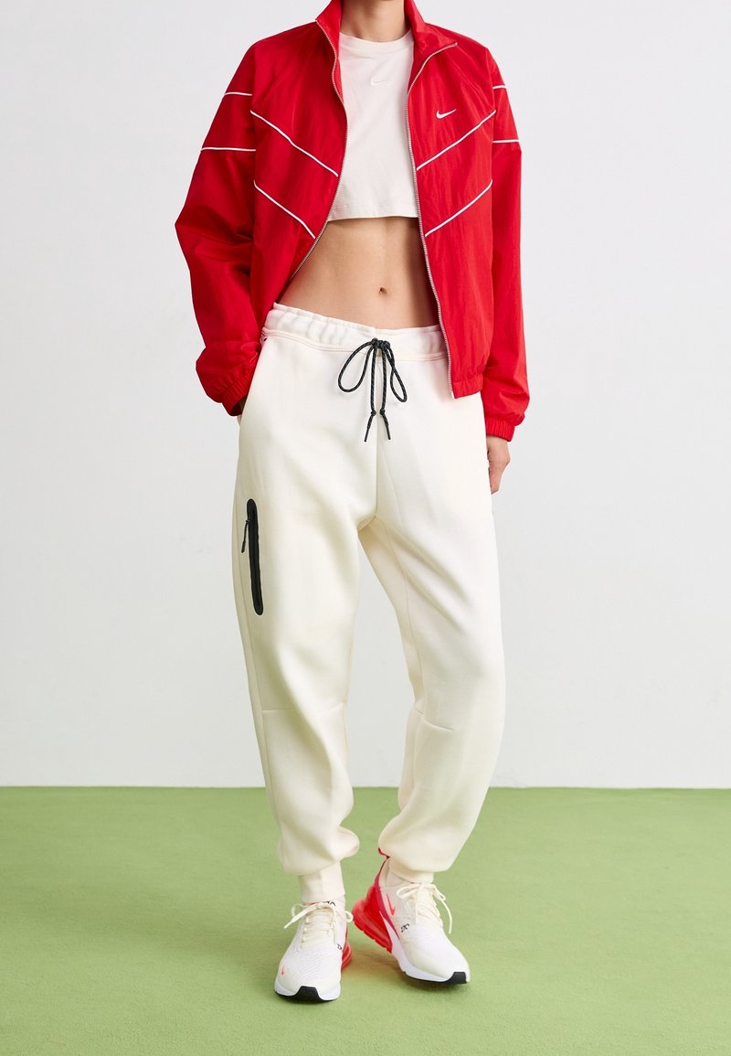 Röd zip-up jacka med vita detaljer, vit kort topp, gräddfärgade sweatpants med svart siddragkedja och vita/röda sportskor.