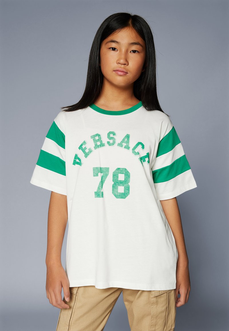 Valge üle mõõtmega t-särk, millel on rohelised triibud varrukatel, kandab tekstuurset rohelise kirjaga "VERSACE" ja "78". Ümmargune kaelusdisain.