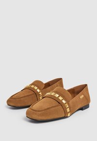 Tanssuede loafers, joissa on litteä korko, koristeellinen nauha kultaisilla niiteillä ja tikatut yksityiskohdat varpaiden alueella. Neutraalit värit.