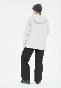 Veste imperméable gris clair avec capuche, associée à un pantalon noir et des chaussures sombres. Texture lisse, design minimaliste, style fonctionnel.