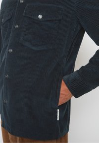 Granatowa koszula ze sztruksu z pionowym prążkowaniem, dwoma kieszeniami na piersi, srebrnymi guzikami i boczną metką. Teksturowany materiał, luźny krój.