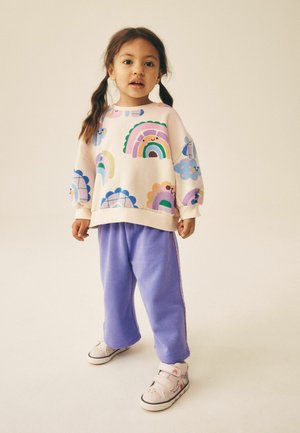 Ung pige med brunt hår i fletninger iført en farverig pastel sweatshirt med regnbuer og blomster, lilla bukser og hvide velcrosko.