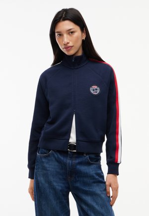 Kvinde iført en marineblå jakke med lynlås, med røde og hvide striber på ærmerne og broderet våbenskjold, kombineret med blå jeans og sort bælte.