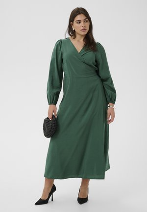 Moteris vilki ilgą žalią suveržiamą suknelę su pūstomis rankovėmis, laiko mažą juodą pintą vokelį ir dėvi juodus aukštakulnius ant balto fono.