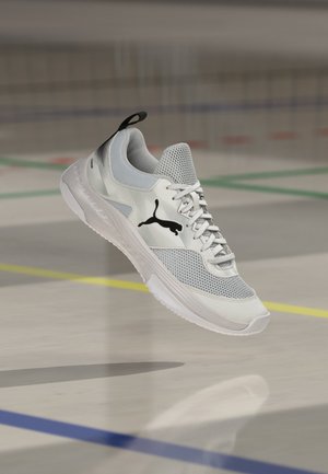 Chaussure de course Puma blanche et grise avec logo noir, flottant au-dessus d’un sol de gym marqué de lignes colorées.