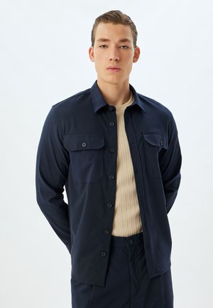 Camisa de botones navy con dos bolsillos en el pecho, hecha de una tela suave, con un corte recto y cuello, llevada sobre una camiseta beis de canalé.