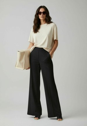 Femme portant un t-shirt beige, un pantalon large noir, des talons ouverts noirs, des lunettes de soleil, et tenant un grand sac rectangulaire beige.