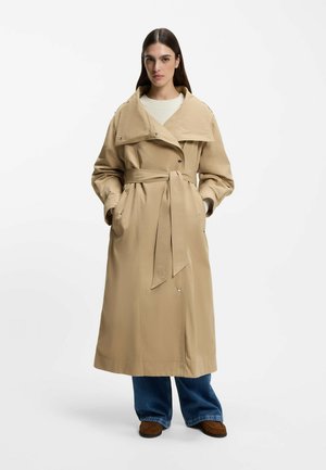 Frau trägt einen beigen Trenchcoat mit Gürtel über einem weißen Pullover, blauen Jeans und braunen Schuhen, steht mit den Händen in den Manteltaschen.