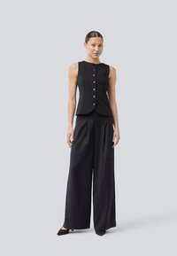 Zwarte mouwloze top met een strakke pasvorm en een knoopsluiting aan de voorkant, gecombineerd met brede zwarte broek, beide in een gladde stof.