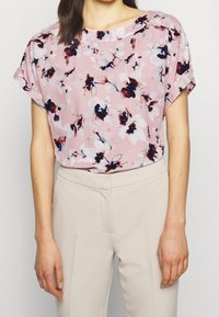 Femme portant un blouse courte à fleurs roses rentrée dans un pantalon taille haute beige, se tenant devant un fond uni.