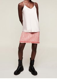 Osoba stojí oblečená v lehkém topu na tenkých ramínkách a červené sukně s vzorem, kterou doplňují černé kozačky po kolena.