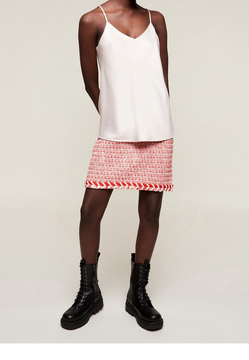 Osoba stojí oblečená v lehkém topu na tenkých ramínkách a červené sukně s vzorem, kterou doplňují černé kozačky po kolena.