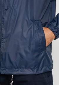 Chaqueta impermeable de color azul marino con textura suave, botones a presión, puños elásticos y un bolsillo lateral. Cuerda en el dobladillo para ajuste.