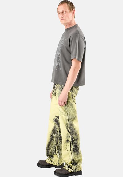 Gray short-sleeve t-shirt with a line drawing design, paired with yellow baggy jeans featuring a distressed graphic pattern and black shoes.