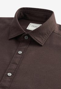 Braunes strukturiertes Hemd mit Button-Down-Kragen, ausgestattet mit zwei grauen Knöpfen und einem Etikett mit der Aufschrift "NEXT TAILORING."