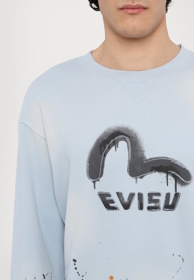 Világoskék hosszú ujjú pulóver, fekete graffitistílusú logóval és festékfröccsökkel. Pamut keverék anyag ribbelt nyakkivágással.