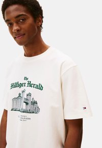 Joven vistiendo una camiseta de manga corta blanca con un paisaje urbano verde y el texto "The Hilfiger Herald" impreso en la parte frontal.