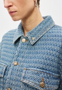 Μπλε τζιν μπουφάν με καπιτονέ ύφασμα, γιακά, χρυσά κουμπιά, δύο μπροστινές τσέπες και ένα μικρό λογότυπο στο γιακά.